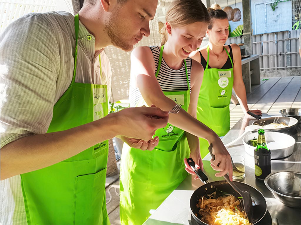 A group in green aprons cooks outdoors, stirring noodles and tasting dishes in a bright, open setting, showcasing teamwork and culinary fun.