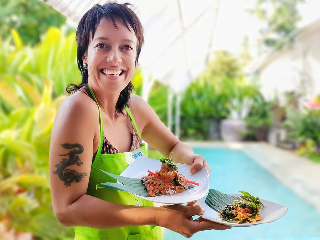 A woman in a green apron proudly holds two plated dishes by a pool, with fresh herbs and vibrant greenery, radiating joy in her cooking success.