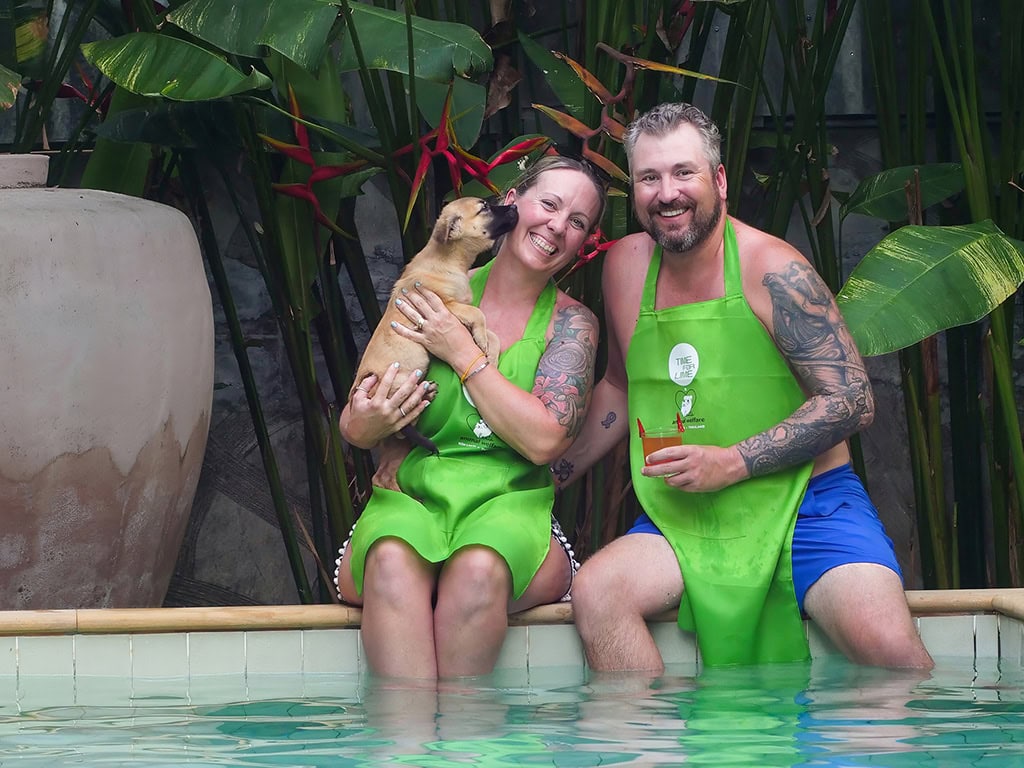 A smiling couple in bright green aprons sits by a pool, with a playful puppy licking the woman’s face. The man holds a drink, and tropical plants surround them.