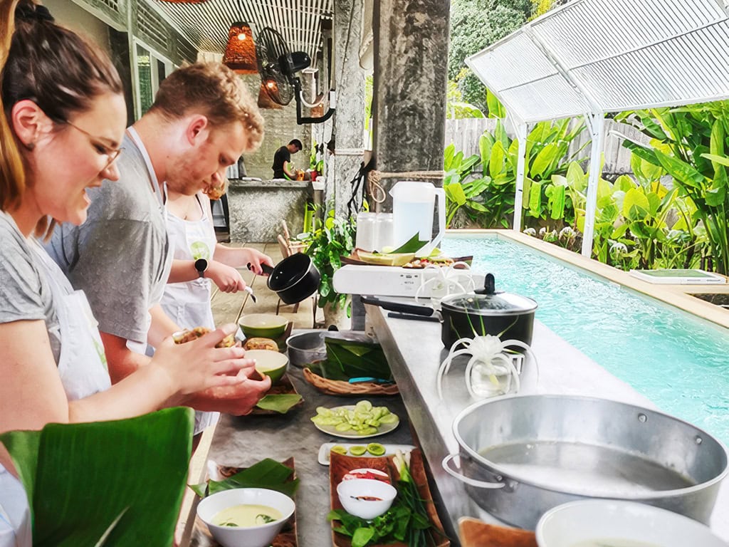 Group of smiling tourists preparing Thai dishes at a cooking station with a pool-side view.